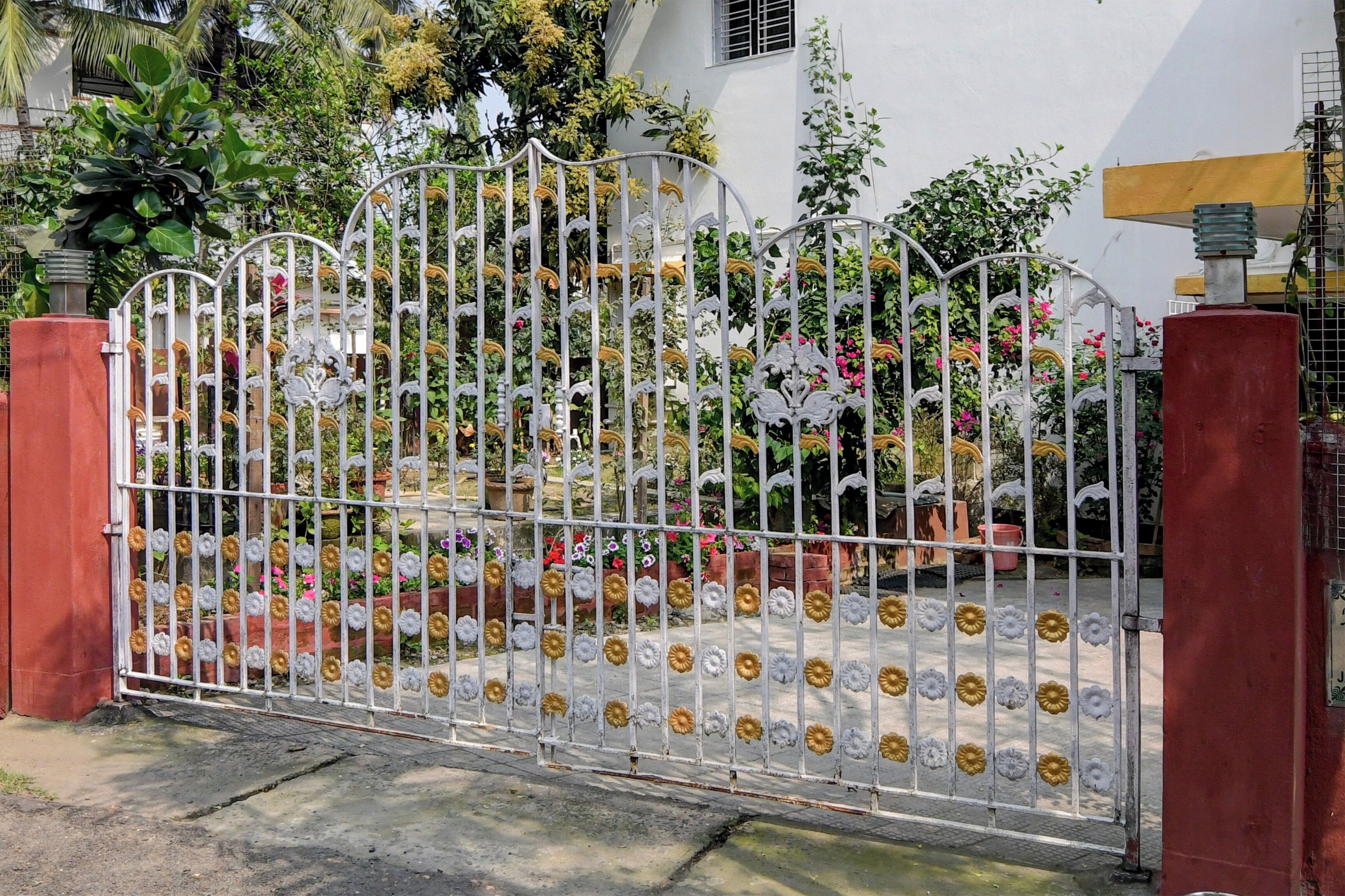 Una entrada con puerta y detalles de metal decorativo y diseños florales, que conduce a un edificio de fachada blanca y abundante vegetación.