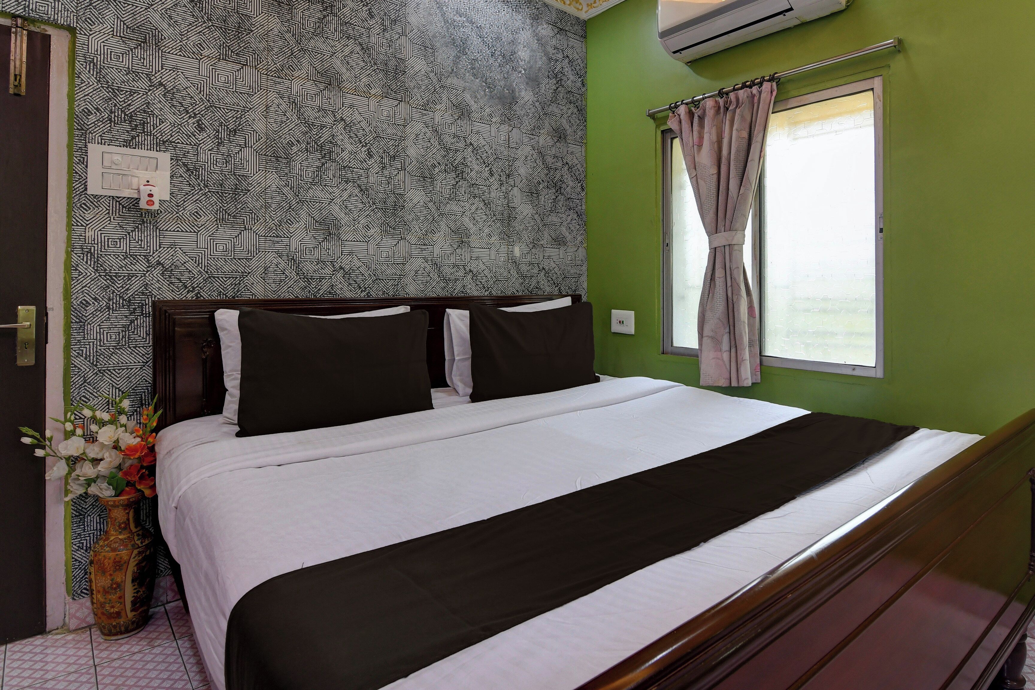 Habitación de hotel con papel tapiz estampado, cama con ropa de cama blanco y negro, ventana con cortinas y un florero con flores.