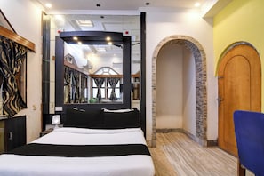 Ett hotellrum med en stor säng, en spegel, en trädörr och en valvformad öppning i sten.