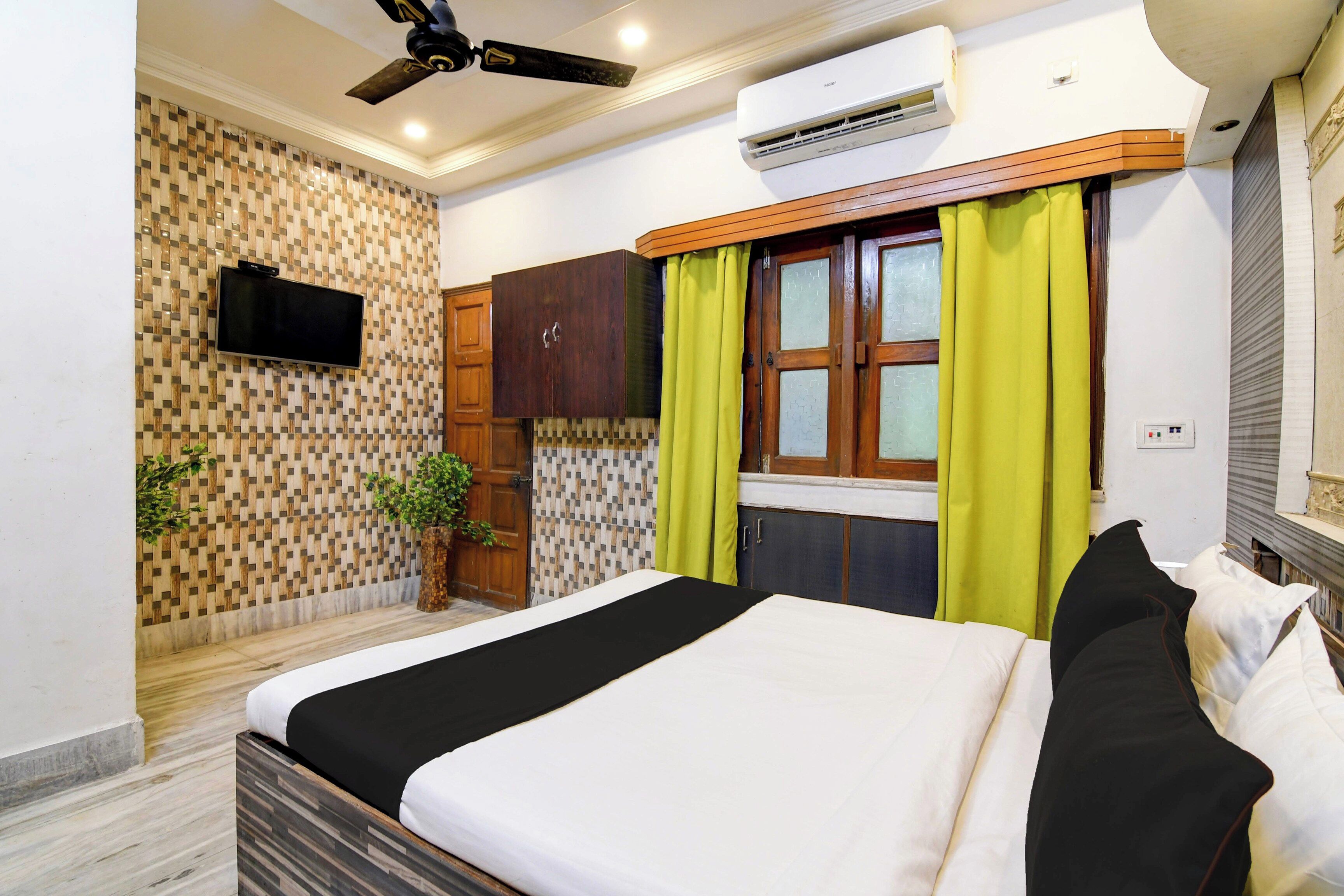 A hotel room with a bed, a television, an air conditioning unit, and a window with curtains.