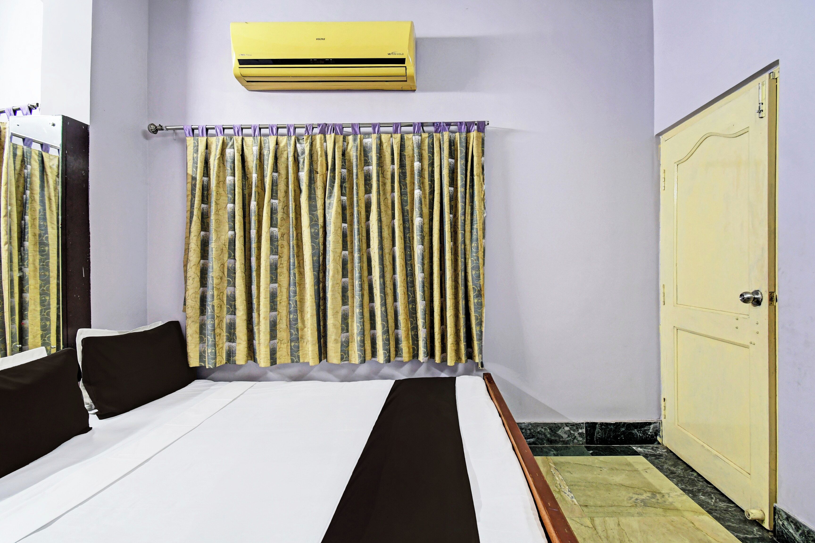 A hotel room with a bed, a wardrobe, an air conditioning unit, and a curtain with a pattern.