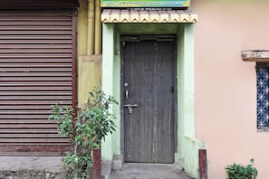 A building with a closed metal shutter door, a wooden door with a latch, and a small window with a grid pattern. - Hotel O Arati House (Howrah)