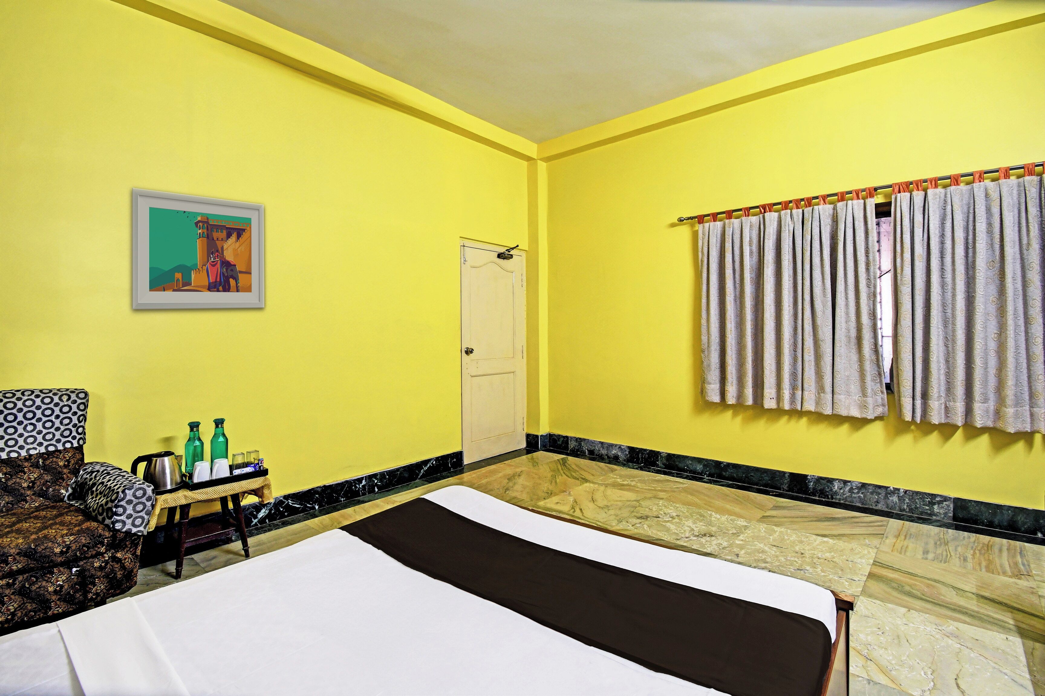 Una habitación con una cama, una mesita de noche con botellas, una silla, una puerta y una ventana con cortinas.