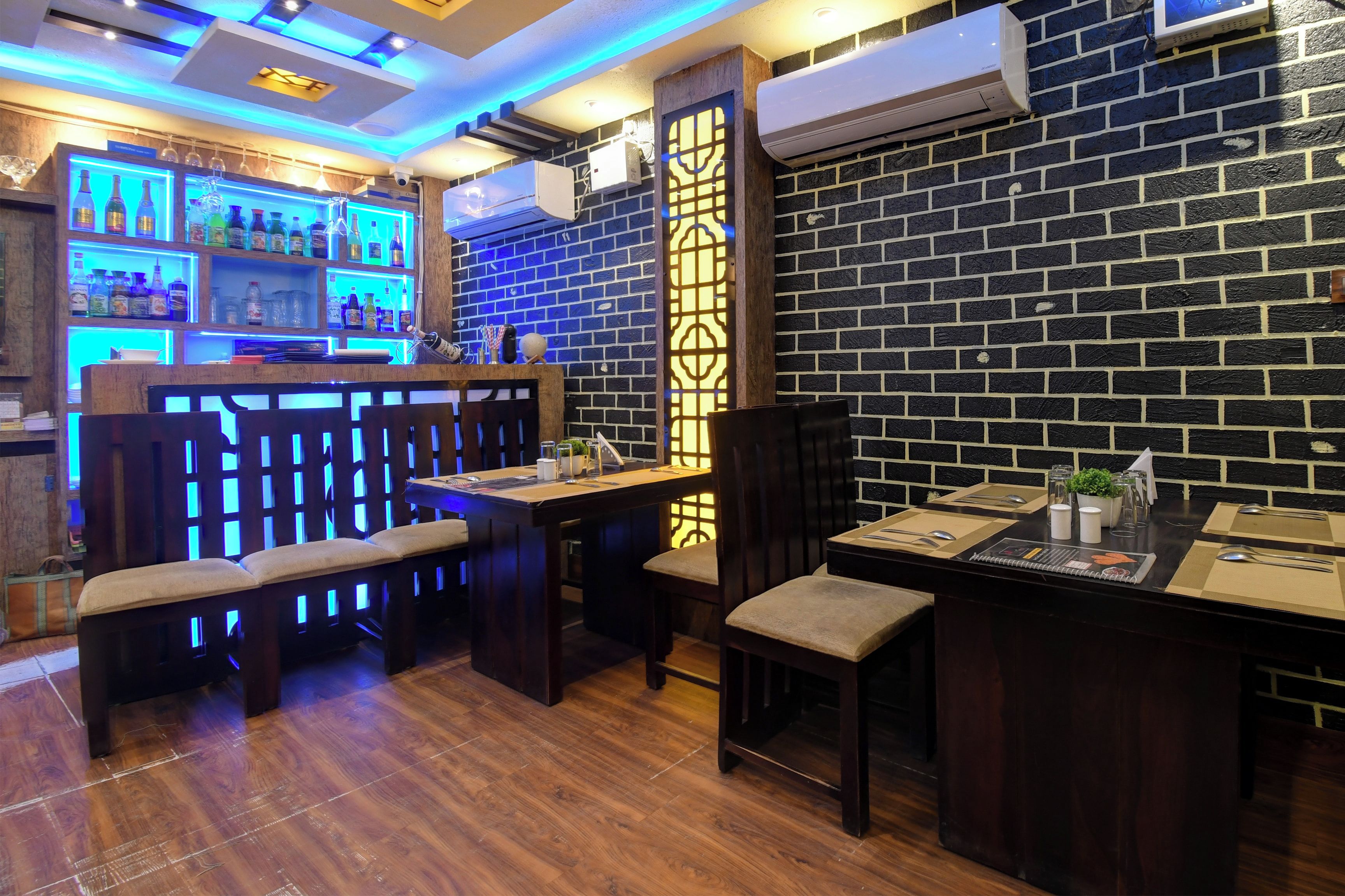 A restaurant interior with wooden furniture, a bar area with shelves of bottles, and a brick wall.