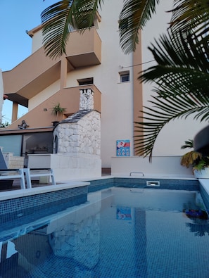 A swimming pool with a stone structure, palm trees, and a building with a balcony. - Villa A GRAND Poolside Apartment  heating pool (Okrug Gornji)