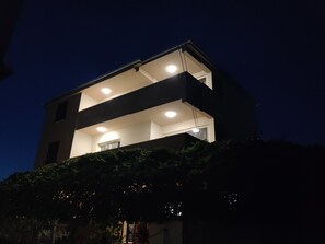 Ein zweistöckiges Gebäude mit Balkon und mehreren Fenstern, umgeben von Grünpflanzen.