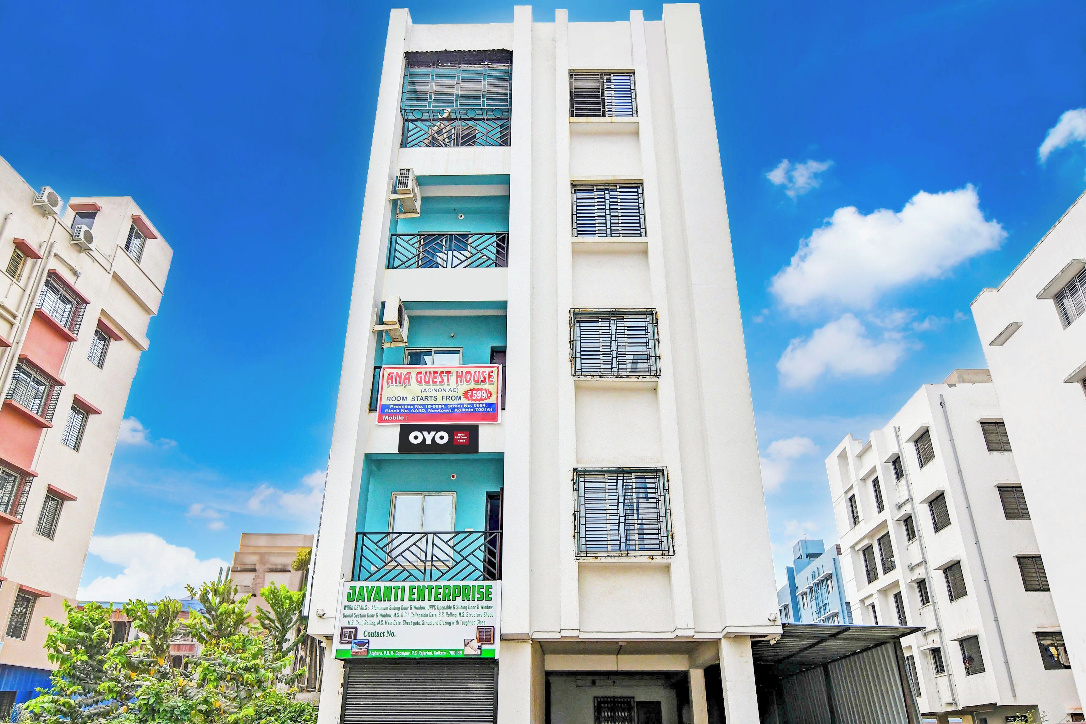 Um edifício de vários andares com varandas, um letreiro de 'ANA GUEST HOUSE' anunciando quartos a partir de ₹599, e outro letreiro de 'JAYANTI ENTERPRISE' detalhando obras no edifício.