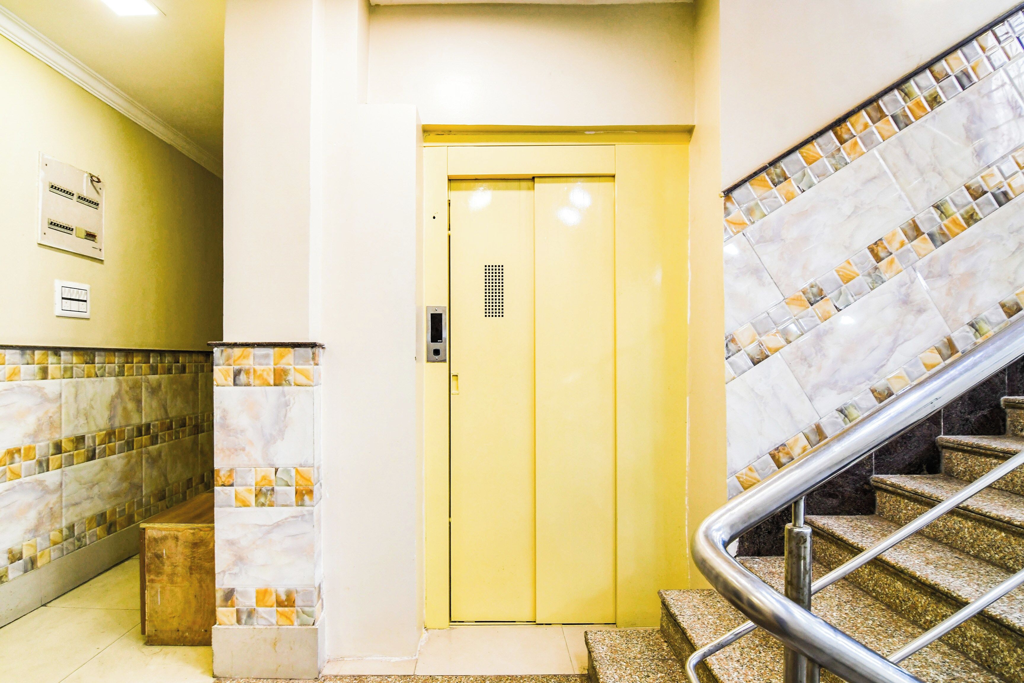 Puerta de ascensor amarilla con un panel de control, pared revestida de azulejos con diseño de mármol y una escalera con barandilla metálica.