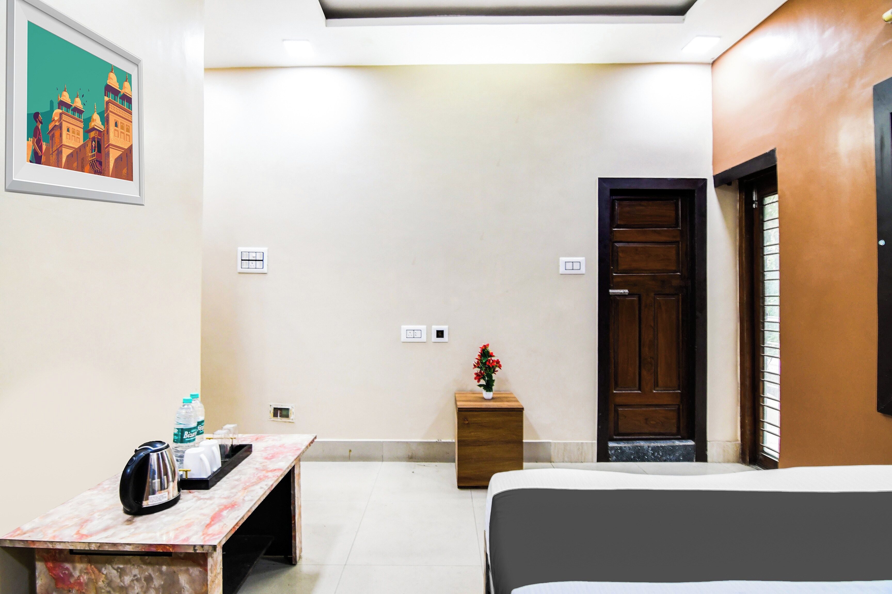 A hotel room with a bed, a small table with a kettle and water bottles, a wooden nightstand with a vase of flowers, and a door with a window.