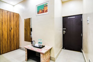 A hotel room with a wooden wardrobe, a small table with a kettle, water bottles, and a framed picture on the wall.