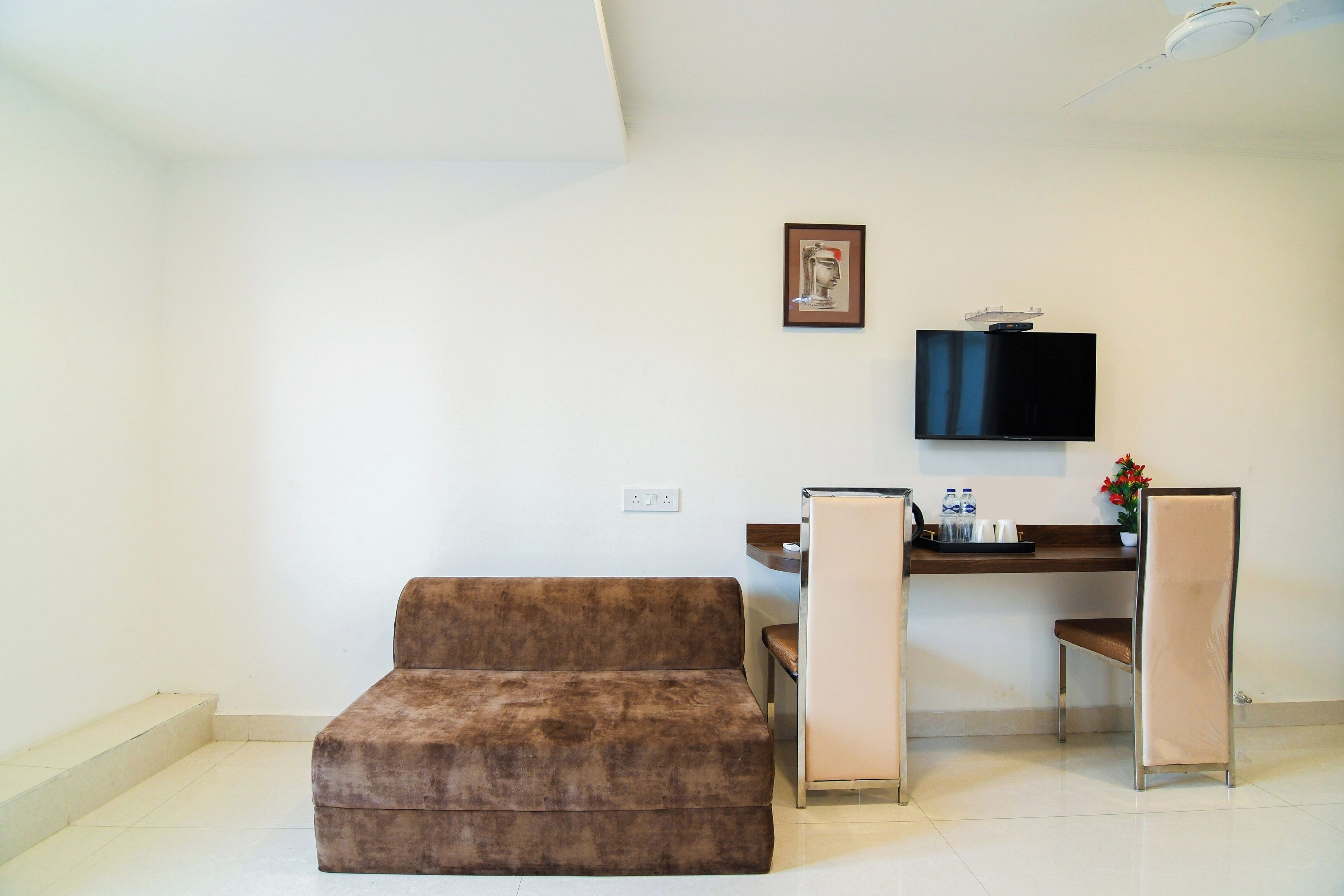 Une chambre meublée comprenant un canapé, un bureau, une chaise, un téléviseur fixé au mur et une petite table sur laquelle est posé un vase de fleurs.