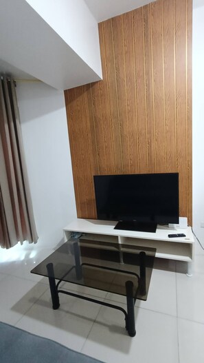A modern living room with a flat-screen TV on a white stand, a glass coffee table, and a wooden panel wall. - Grand Riviera Suite 1BR with 2 beds near SLEC & US EMBASSY (Manila)