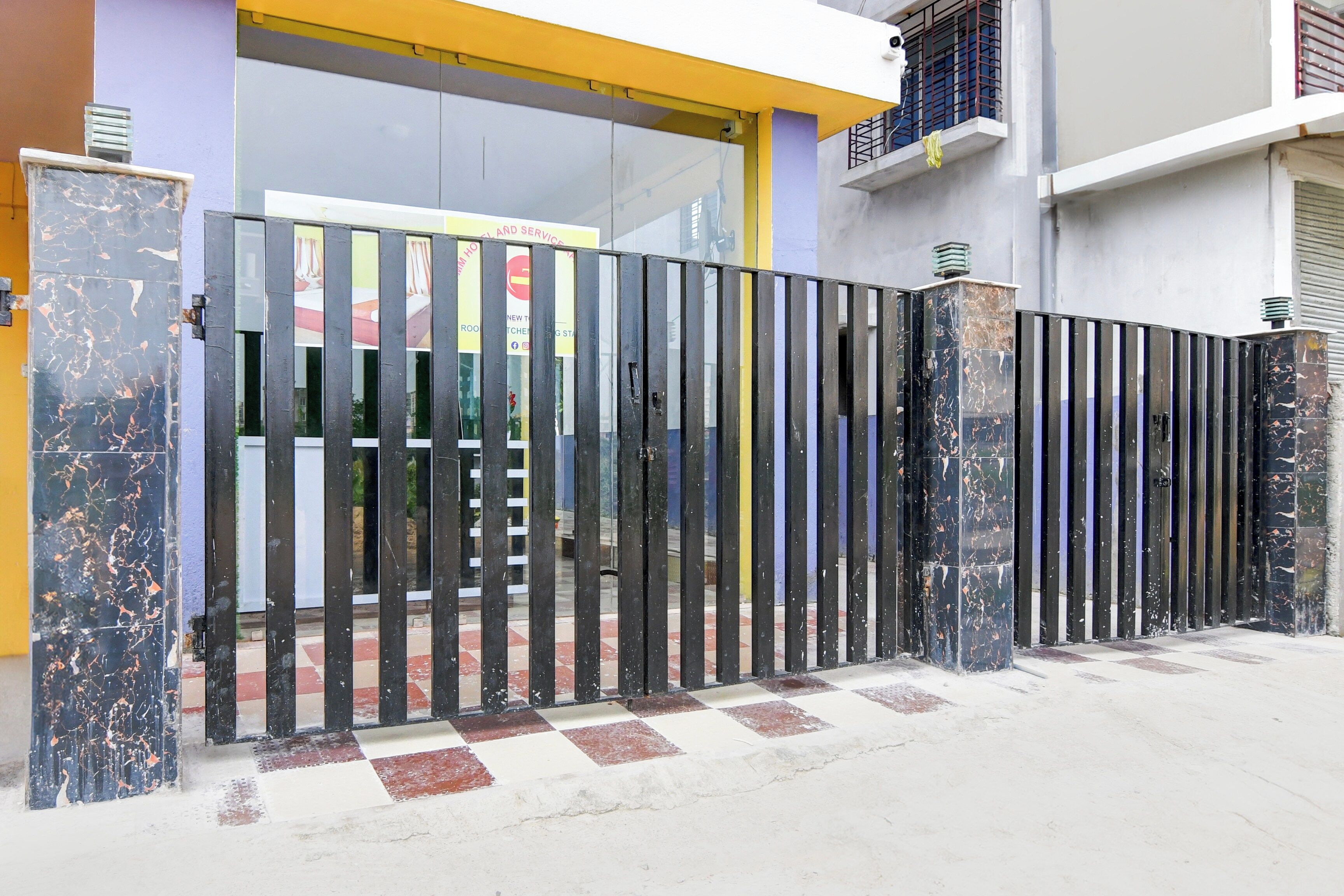 A gated entrance with a checkered floor pattern, a metal gate, and a building with a yellow and grey facade.