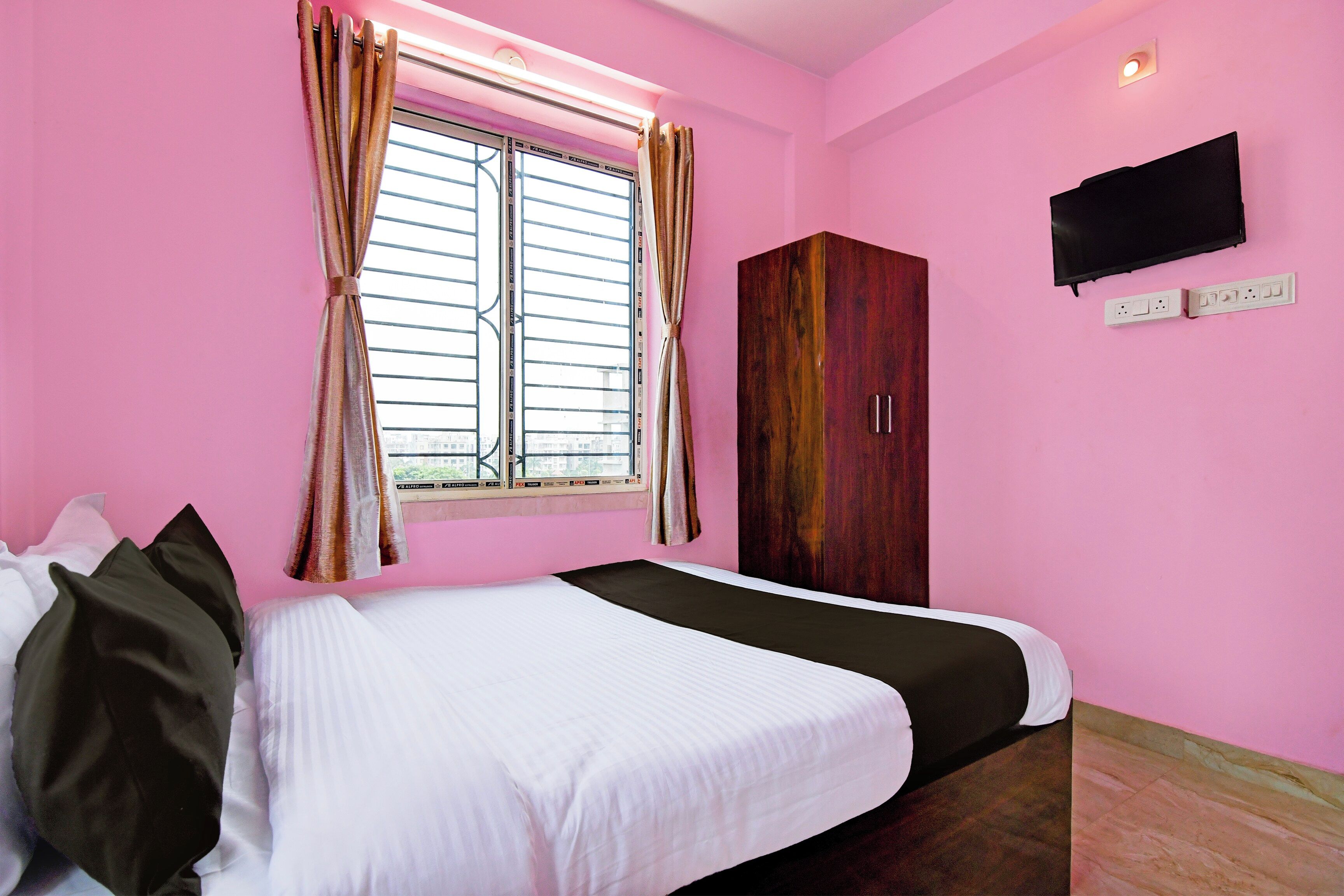 A room with a bed, a wardrobe, a window with curtains, and a flat-screen TV mounted on the wall.