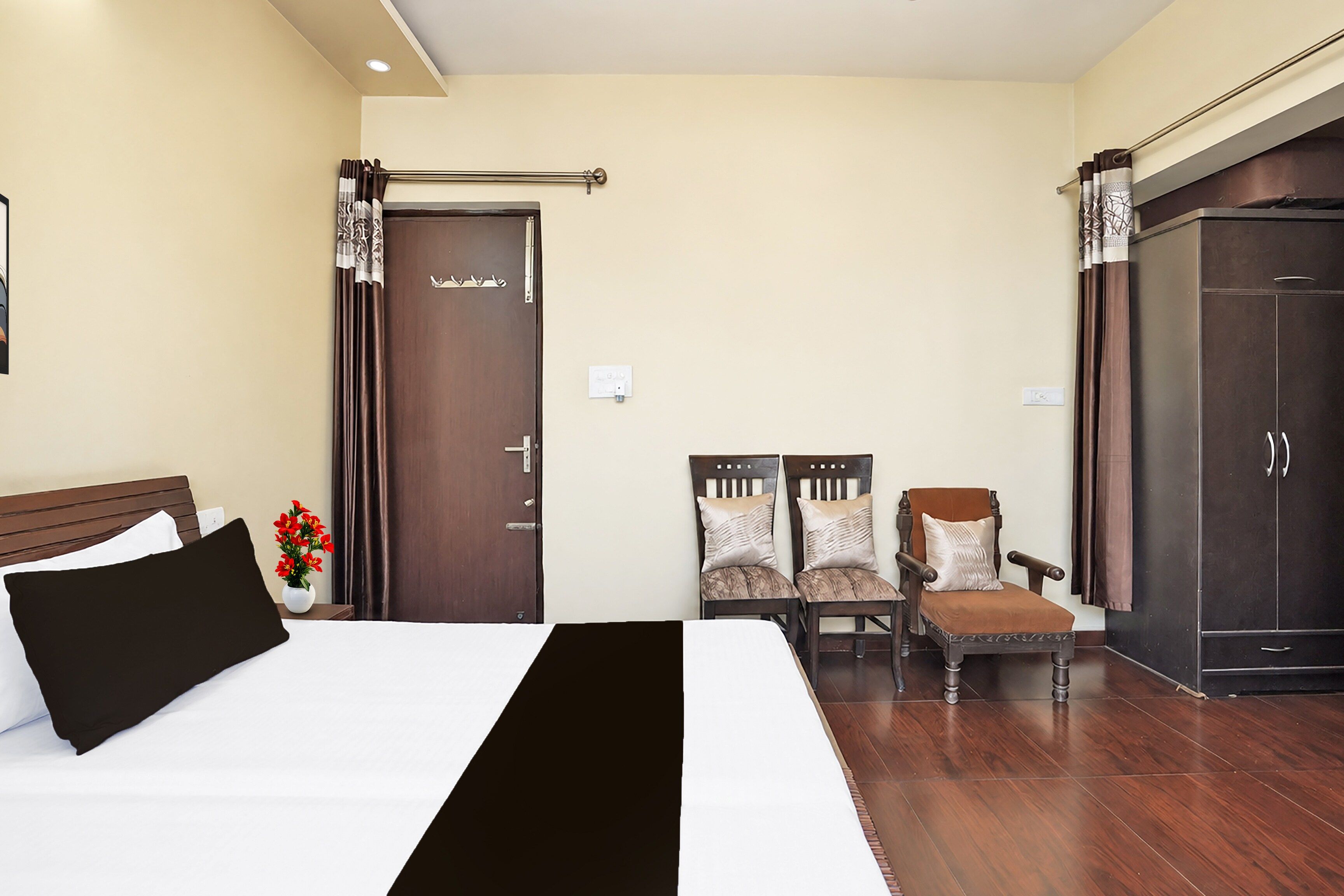A hotel room with a bed, two chairs, a wardrobe, and a small table with a vase of flowers.