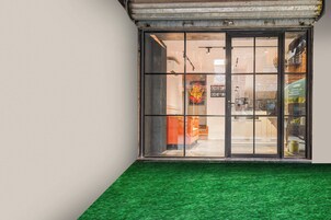 Fachada con un acceso de césped artificial verde, puerta de vidrio y un cartel de recepción/salida, junto con el reflejo de un edificio en el vidrio.