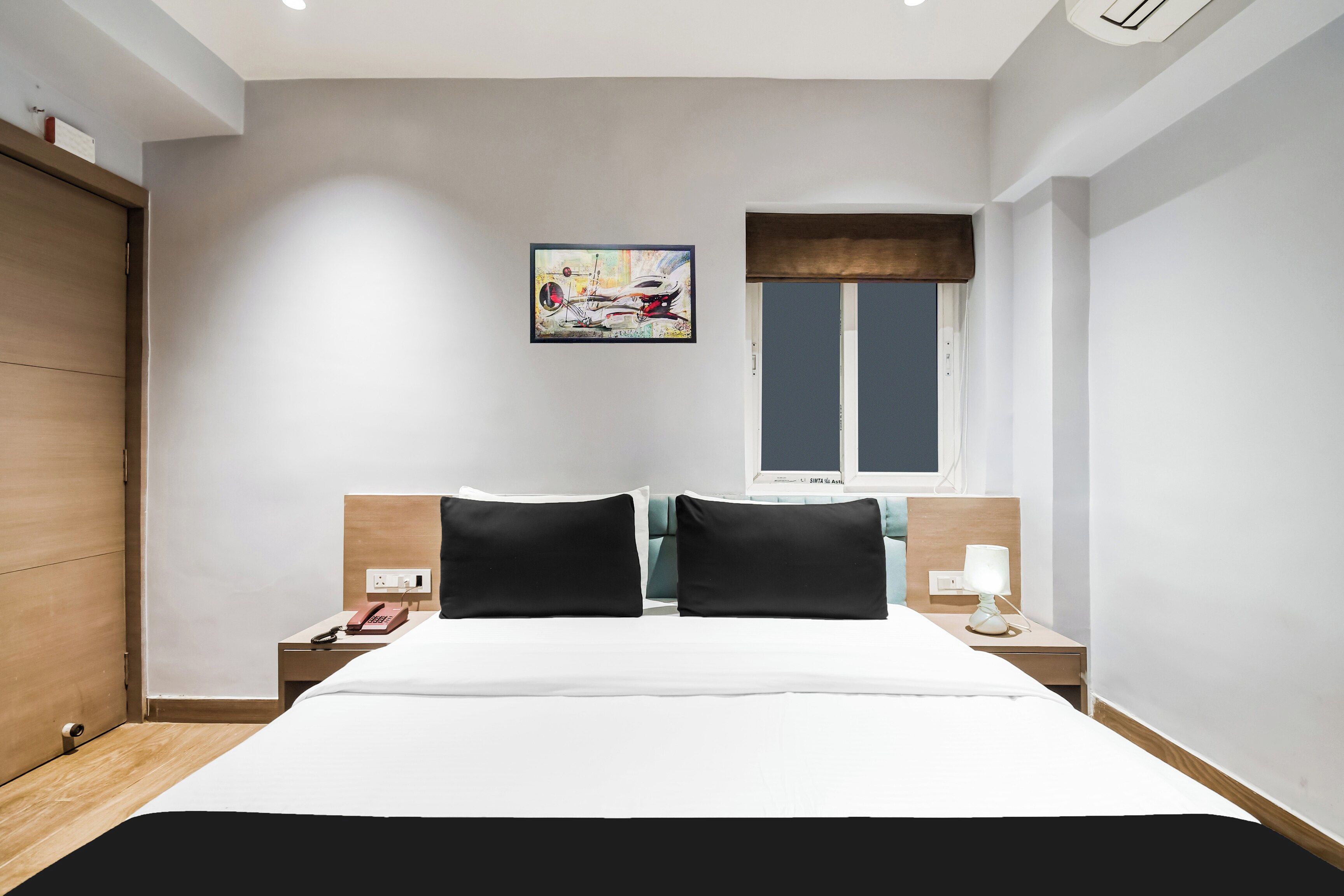 A hotel room with a bed, two bedside tables, a nightstand, a painting on the wall, and a window.