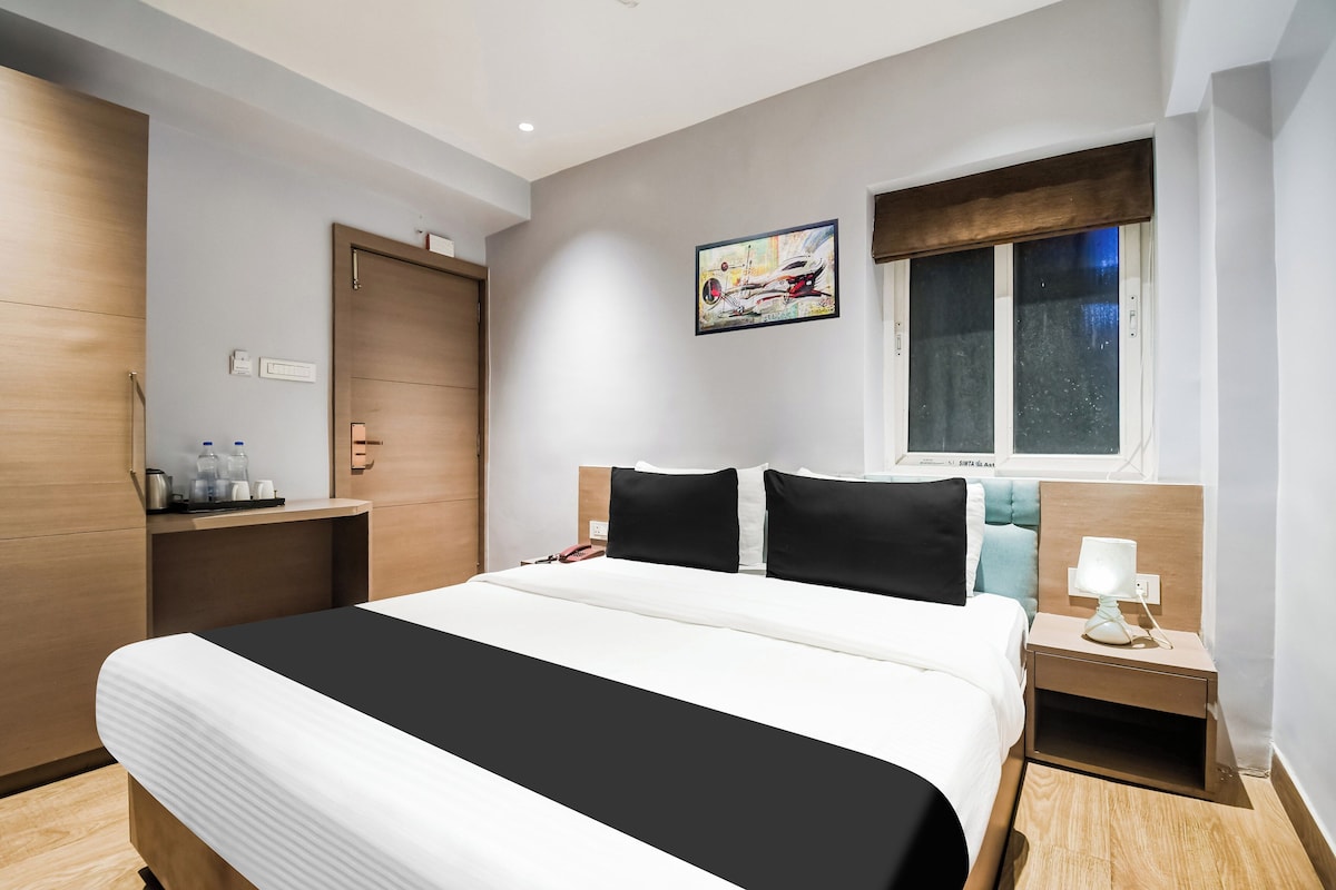 A hotel room with a bed, bedside table, nightstand, window, and artwork on the wall.