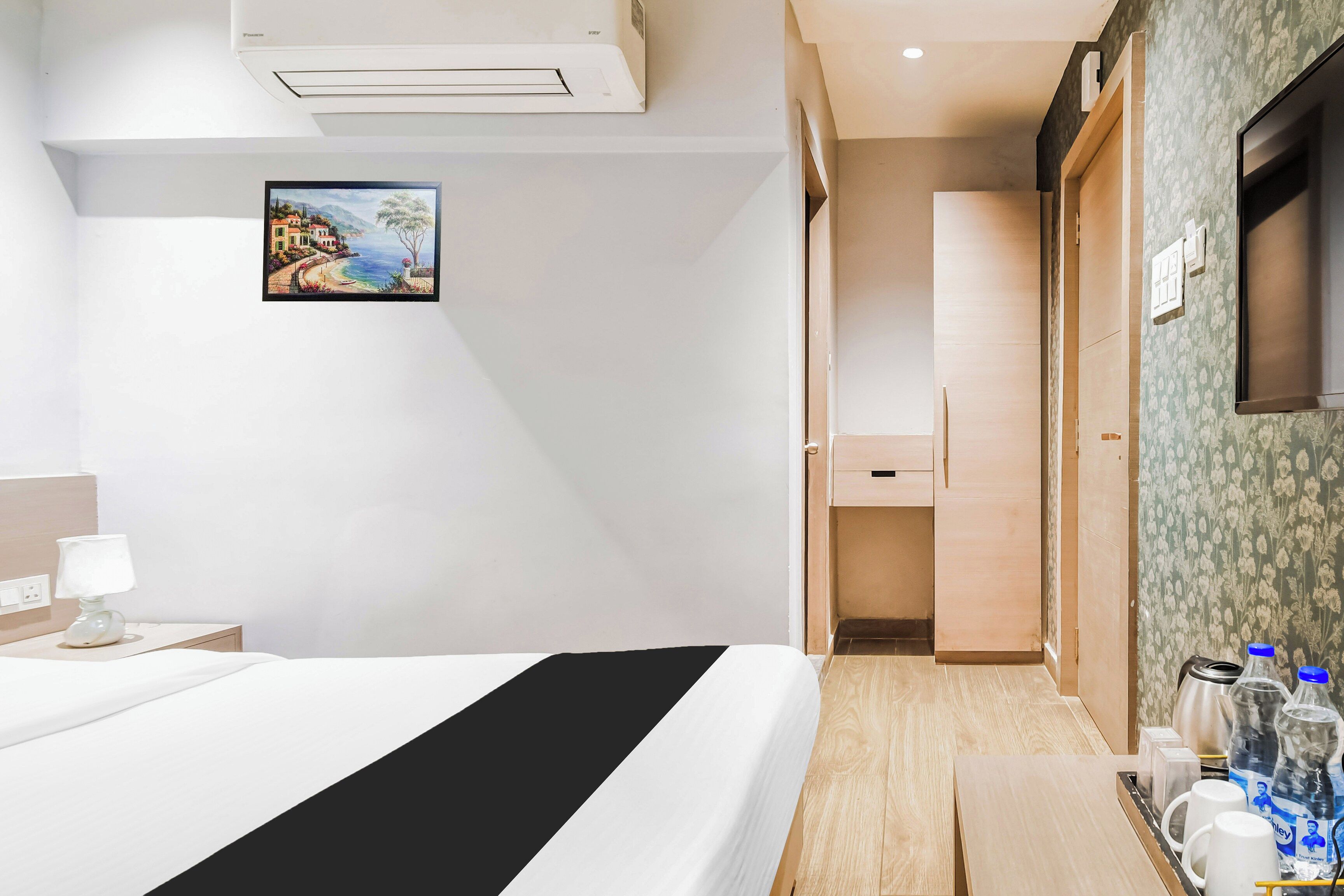 Une chambre d’hôtel avec un lit, une table de chevet avec une lampe, un tableau au mur, une armoire et une salle de bain visible à travers une porte ouverte.