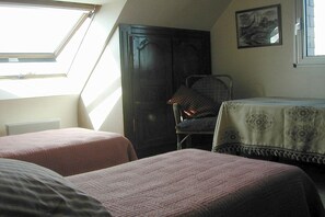 Ein Schlafzimmer mit zwei Betten, einem Sessel, einem Tisch mit Tischdecke, einem dunklen Holzschrank und einem Bild an der Wand.