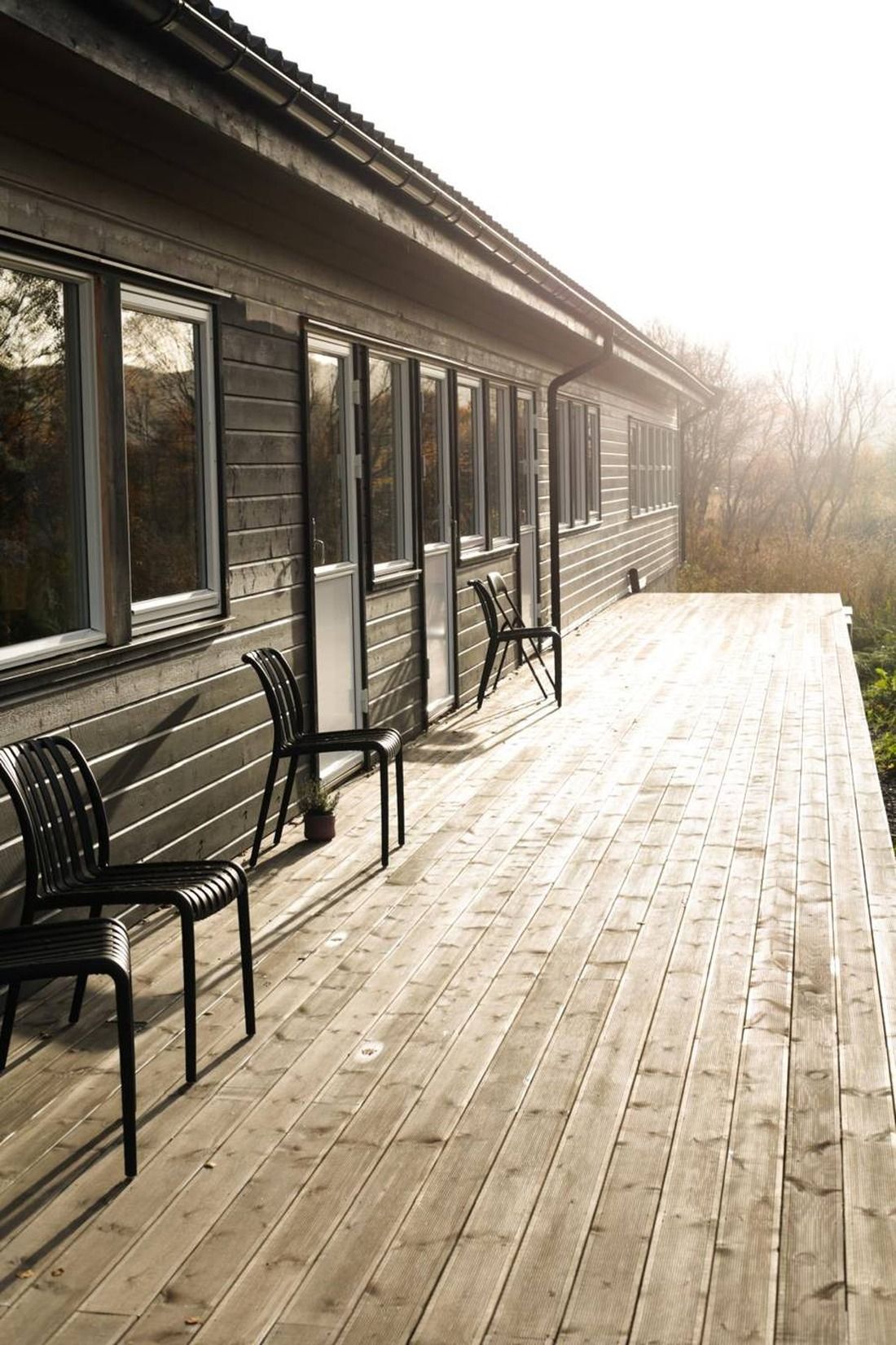 Una terraza de madera con mobiliario exterior y un edificio con ventanas amplias.