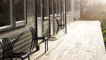 Una terraza de madera con mobiliario exterior y un edificio con ventanas amplias.