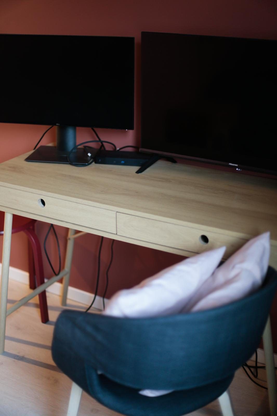 Ein Schreibtisch aus Holz mit zwei Monitoren, einer Tastatur und einer Maus. Davor steht ein Sessel mit Kissen.