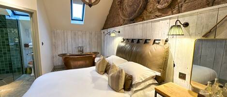 A bedroom with a large bed, a wooden headboard, a stone wall, a bathtub, and a chandelier.