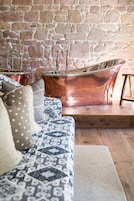 Una habitación con pared de piedra, una bañera de cobre, un sofá estampado y un taburete de madera.