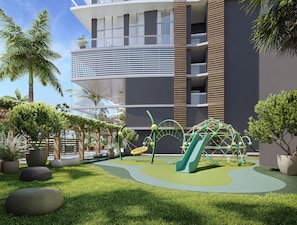 A modern playground with slides and climbing structures, surrounded by grass and potted plants, with a building featuring balconies in the background. - Premier Studio | Brand New | Resort Pool + Gym | Near Beach | Nexo Miami (North Miami Beach)