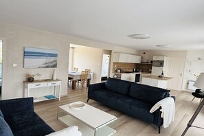 A modern living room with a dark blue sofa, a glass coffee table, and a dining area with a white table and chairs. - Les Mouettes just 50 m from the sea (Plouescat)