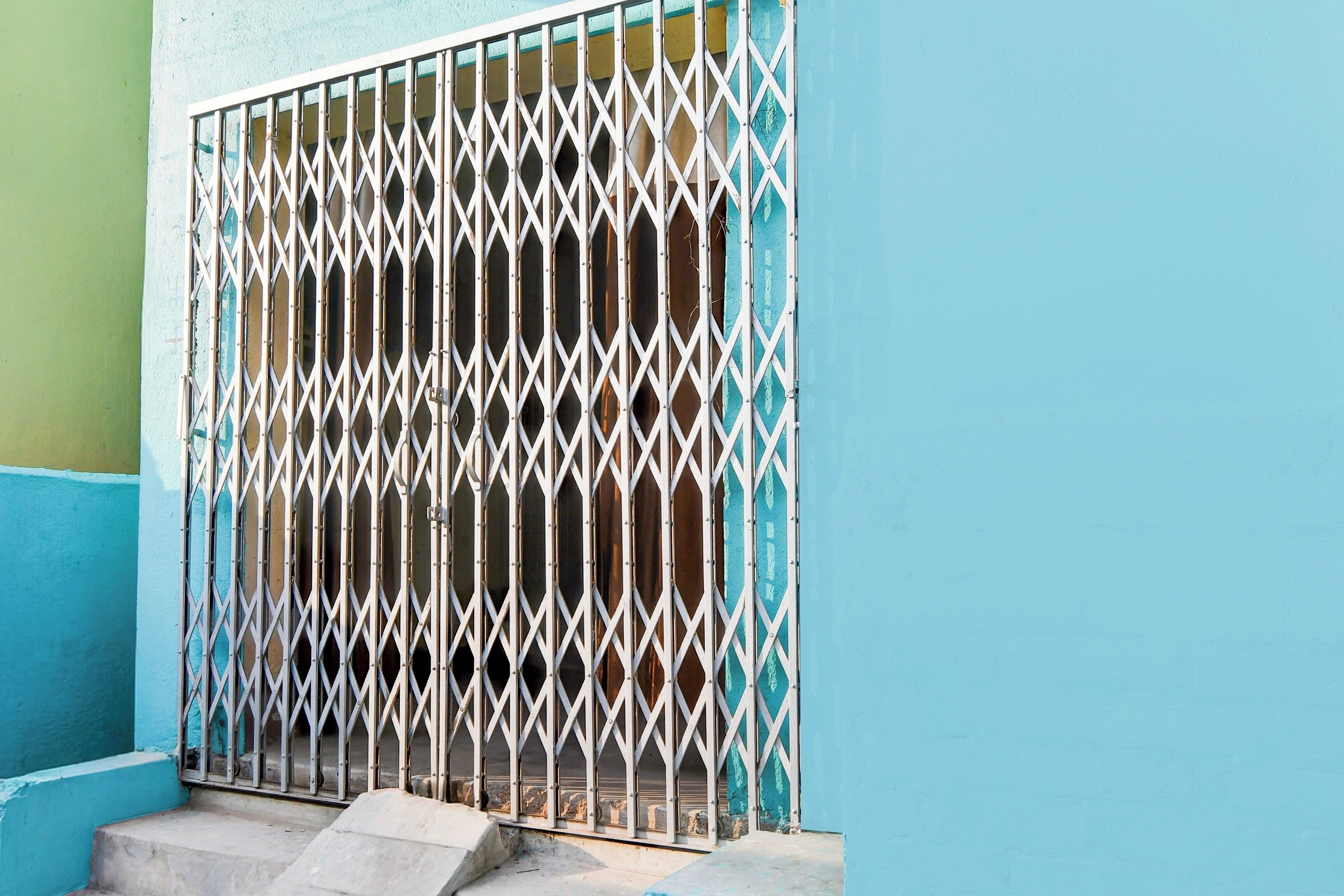 Una verja de metal con diseño de rombos, adosada a un edificio de fachada azul y verde.