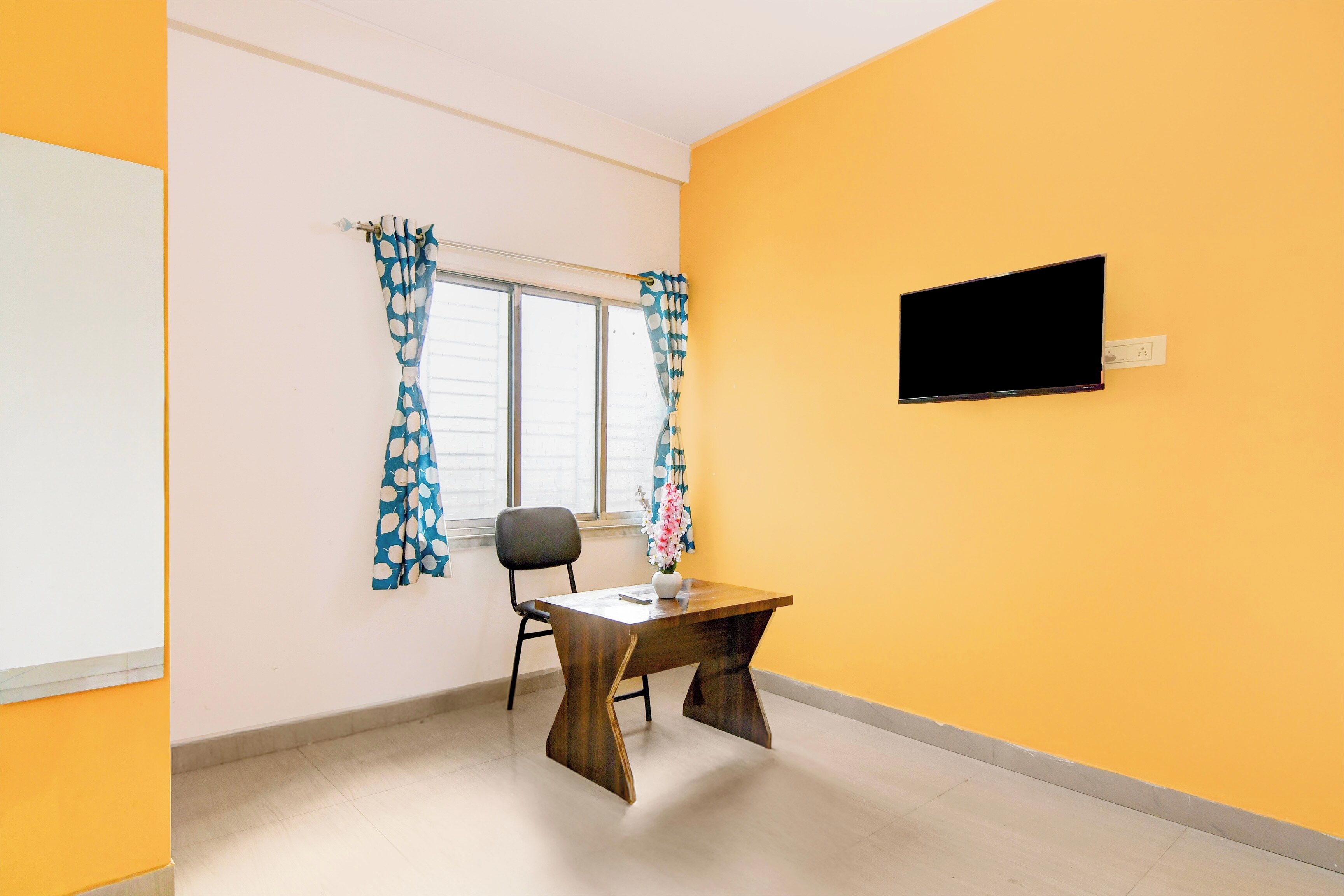 A room with a single table, a chair, a window with curtains, and a mounted television.