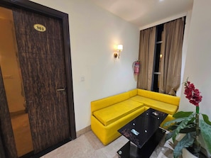 A room with a yellow sofa, a black glass coffee table, a door with a number plate, a wall-mounted light, and a potted plant. - Hotel O cloud 9 (Amritsar)