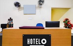 Una recepción de hotel con un letrero que dice 'HOTELO', dos ventiladores de pared, una computadora portátil, un jarrón con flores rojas y una silla azul.