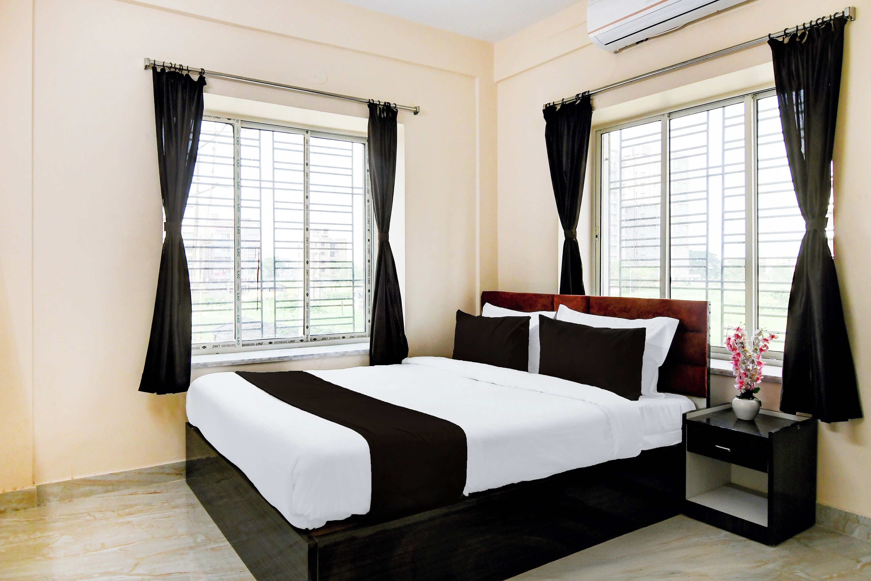 A hotel room with a bed, a nightstand, a vase with flowers, and two windows with curtains.