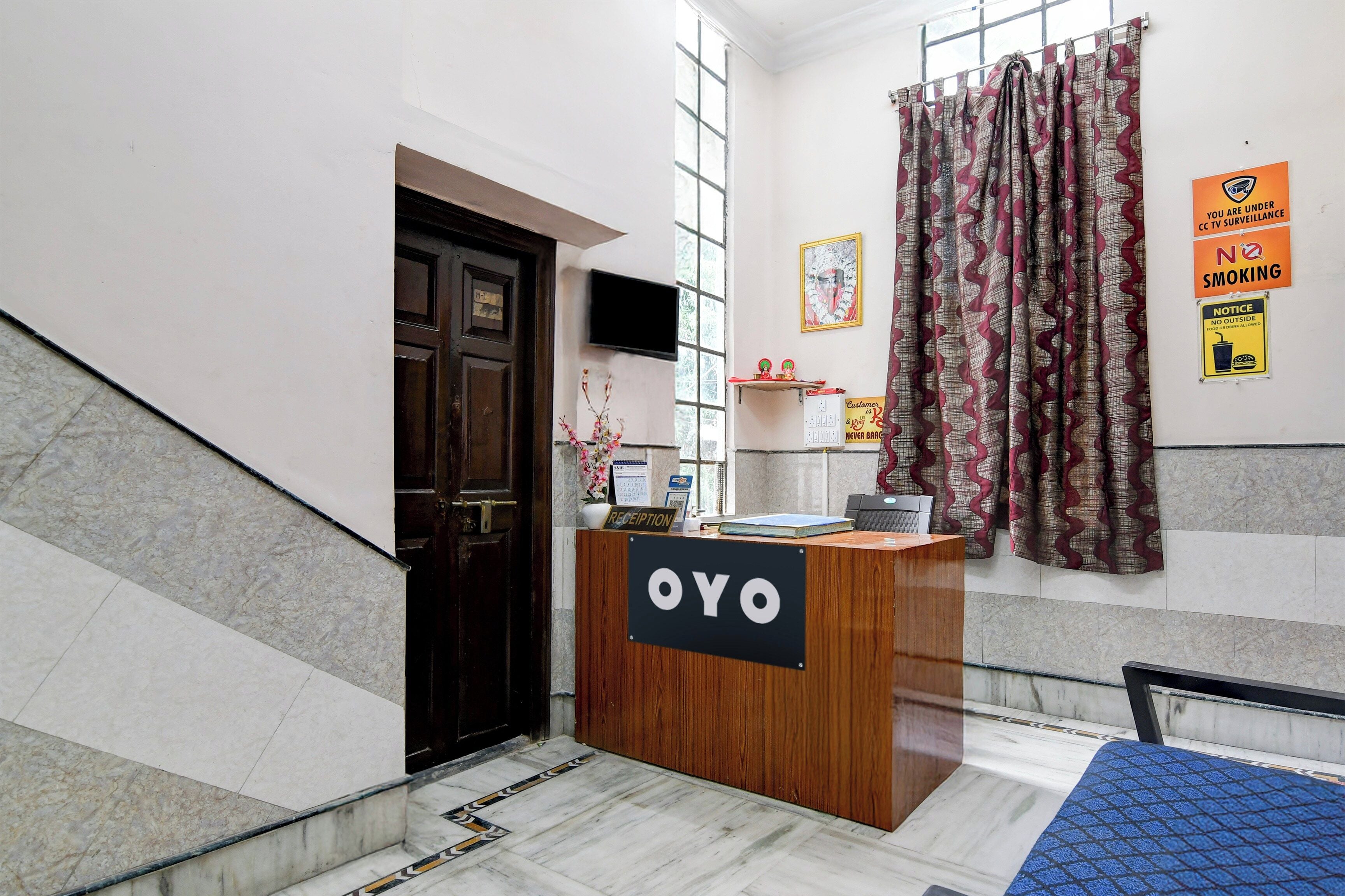 Eine Rezeption mit einem Schild, auf dem 'OYO' steht, in einer Hotelhalle mit Fliesenboden, einer Holztür und einem Fenster mit gemusterten Vorhängen.