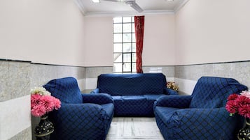 A room with a blue patterned sofa set, a ceiling fan, a window with red curtains, and a vase with flowers.