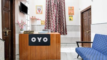 A reception desk labeled 'OYO' with a blue patterned chair, a TV mounted on the wall, and a curtain with a wavy pattern.