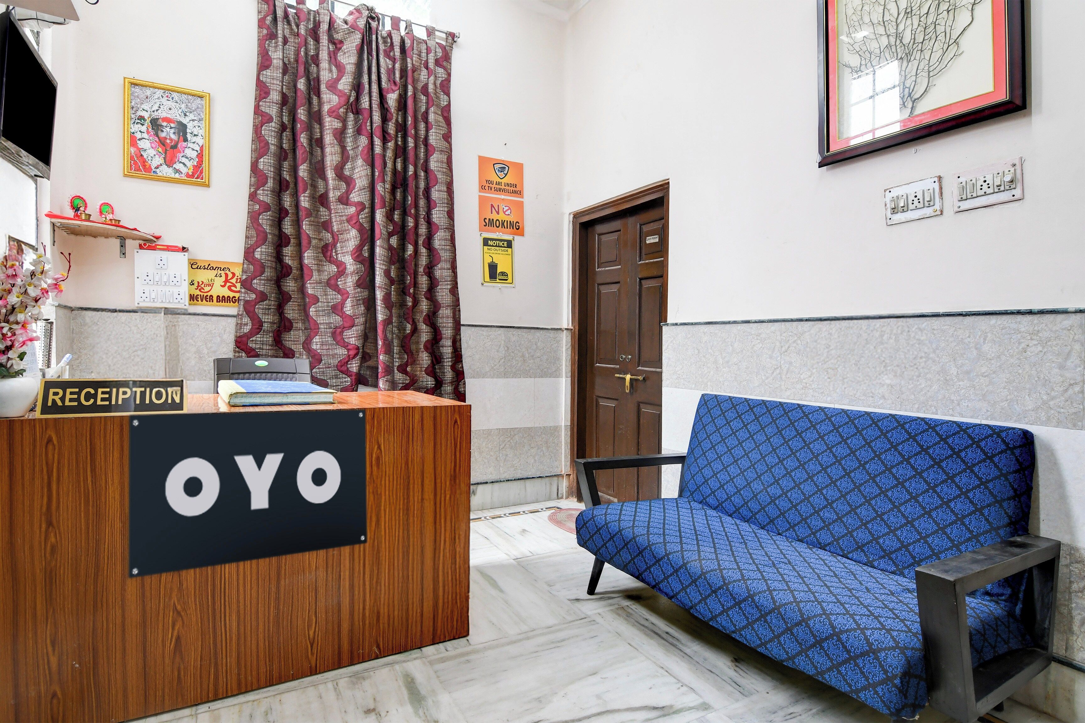 A reception desk with a sign reading 'RECEPTION' and 'OYO'. There is a blue patterned sofa and a door in the background.