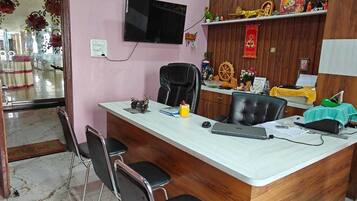 Una mesa de recepción con un televisor de pantalla plana, una computadora y un teléfono. Hay sillas alrededor de la mesa, y un gabinete de madera con objetos decorativos en la pared de fondo.