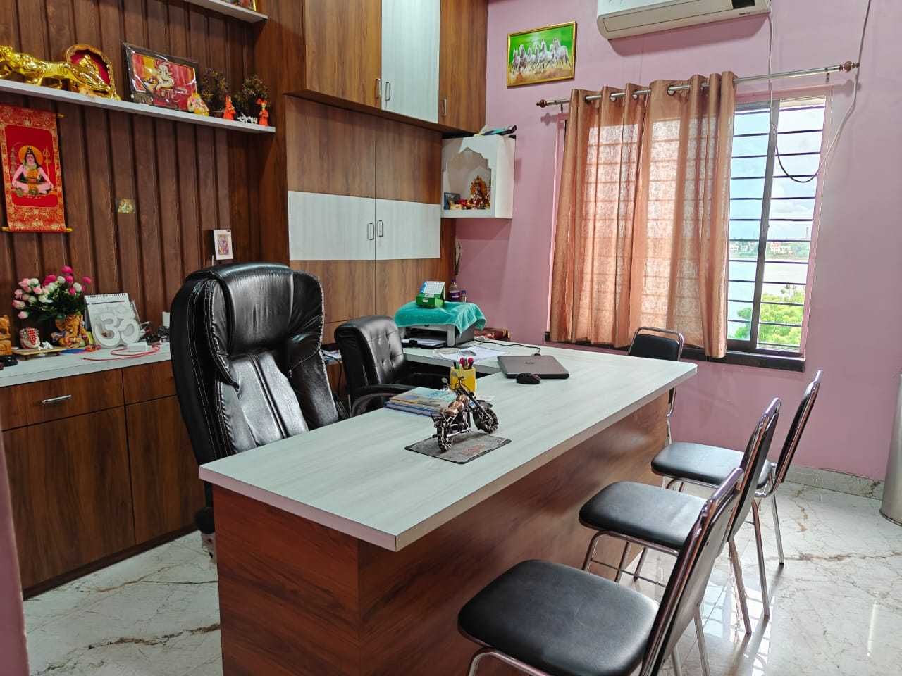 Una habitación con un escritorio de madera, una silla de oficina negra, una pequeña mesa central con cuatro sillas y una ventana con cortinas.