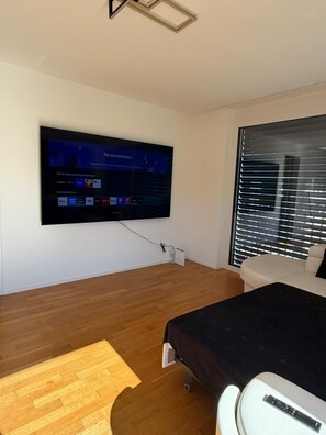 A modern bedroom with a flat-screen TV mounted on the wall, a bed with a black cover, a wooden floor, and a window with blinds.