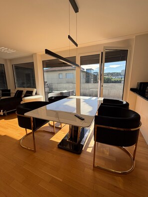 A modern dining area with a glass table, black chairs, and a view of the city through large windows.