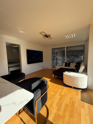 A modern living room with a flat-screen TV mounted on the wall, a sofa, a coffee table, and a dining area with a marble countertop.
