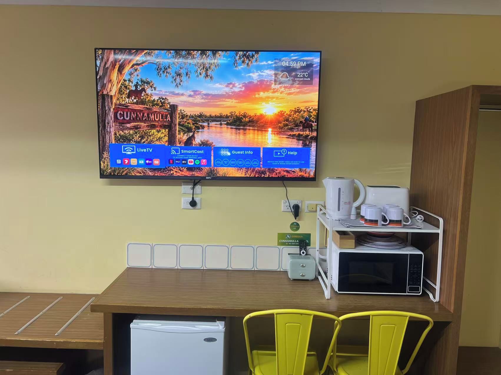 Un televisor montado en la pared que muestra un atardecer panorámico, una cafetera, un microondas, un refrigerador pequeño y una mesa con sillas.