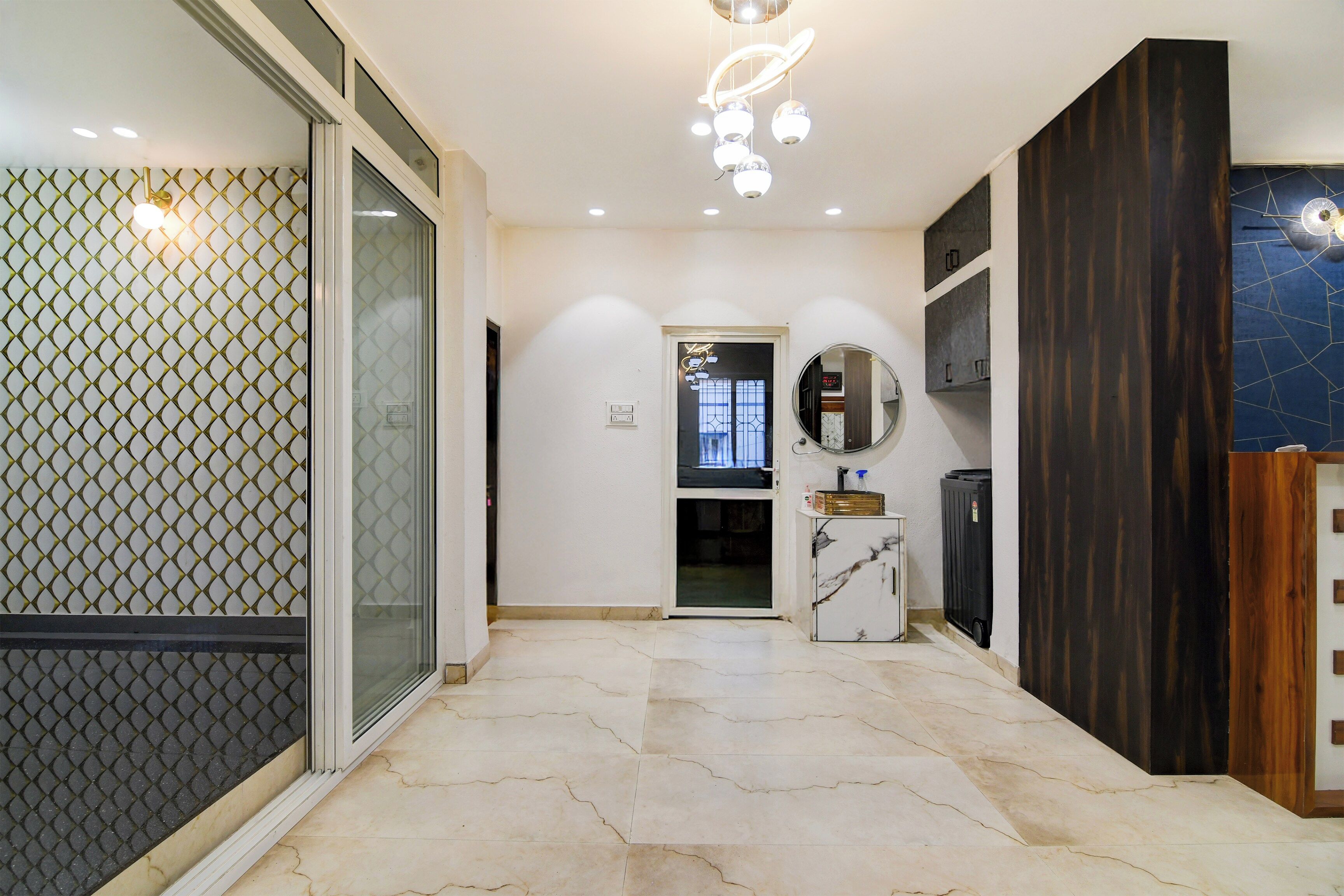 A hallway with a glass door, a marble floor, a dark wooden panel, and a round mirror.
