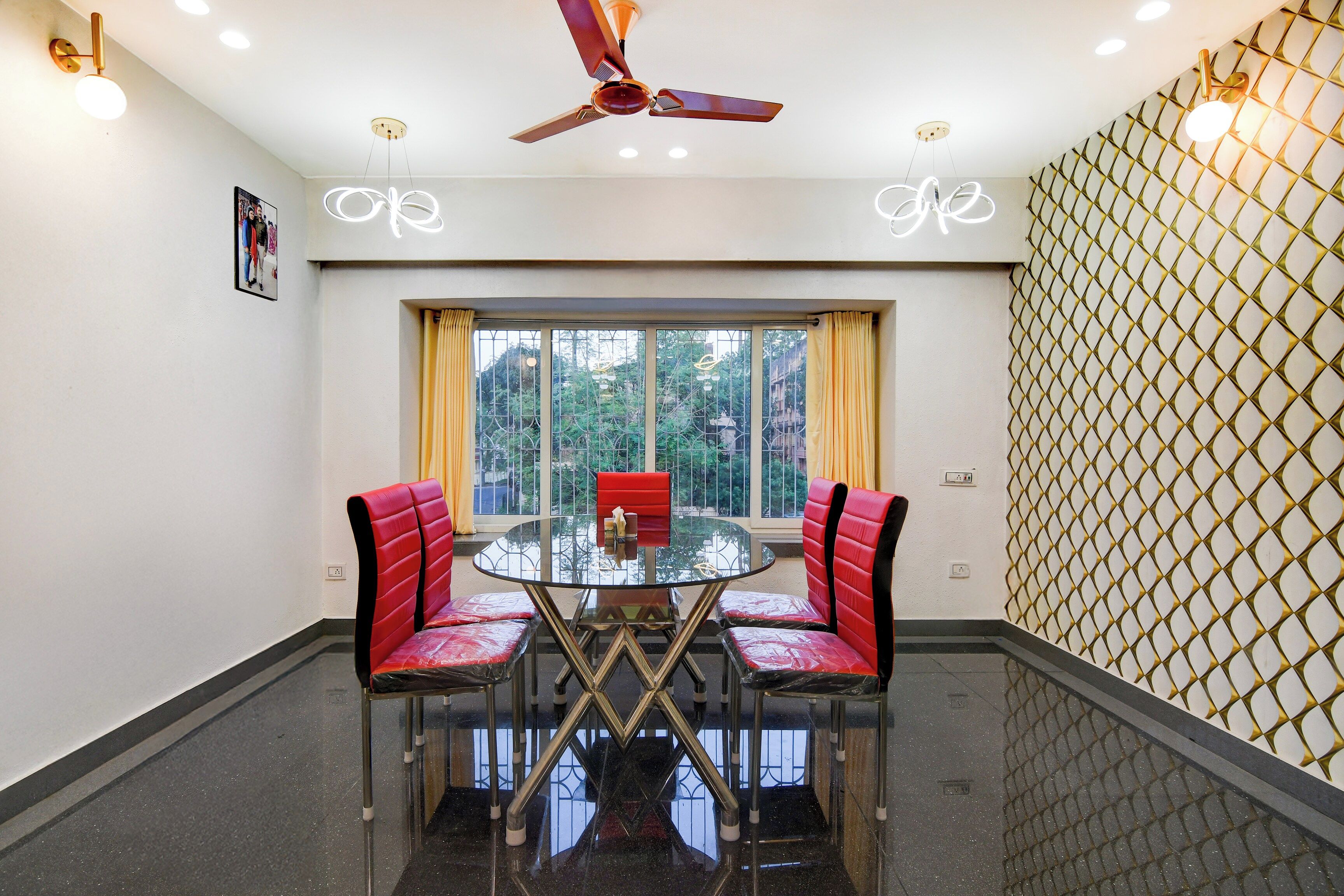 A room with a round glass table, two red upholstered chairs, a ceiling fan, and a window with curtains.