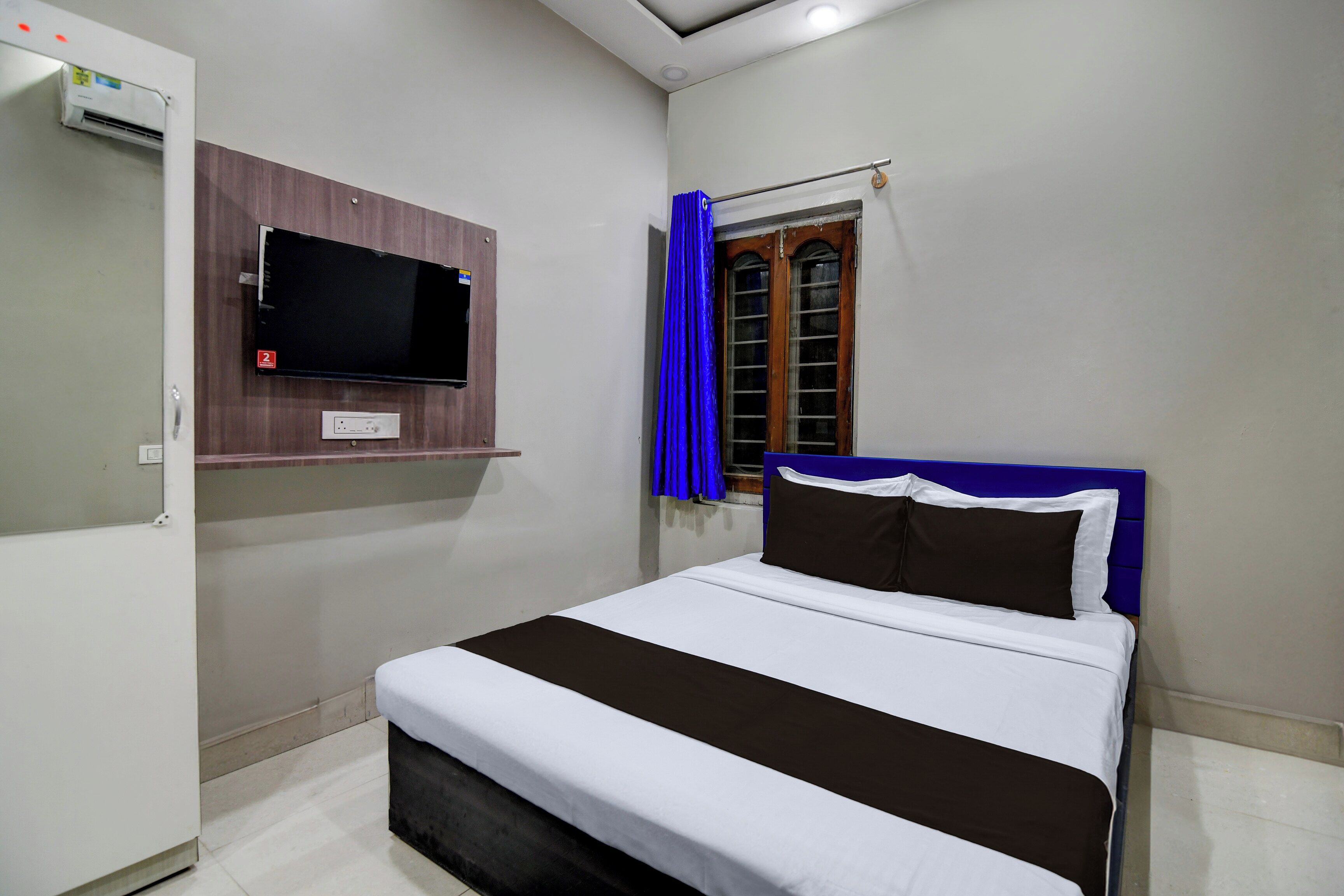Ein Schlafzimmer mit einem Bett, einem an der Wand befestigten Fernseher, einem Fenster mit Vorhängen und einem Kleiderschrank.