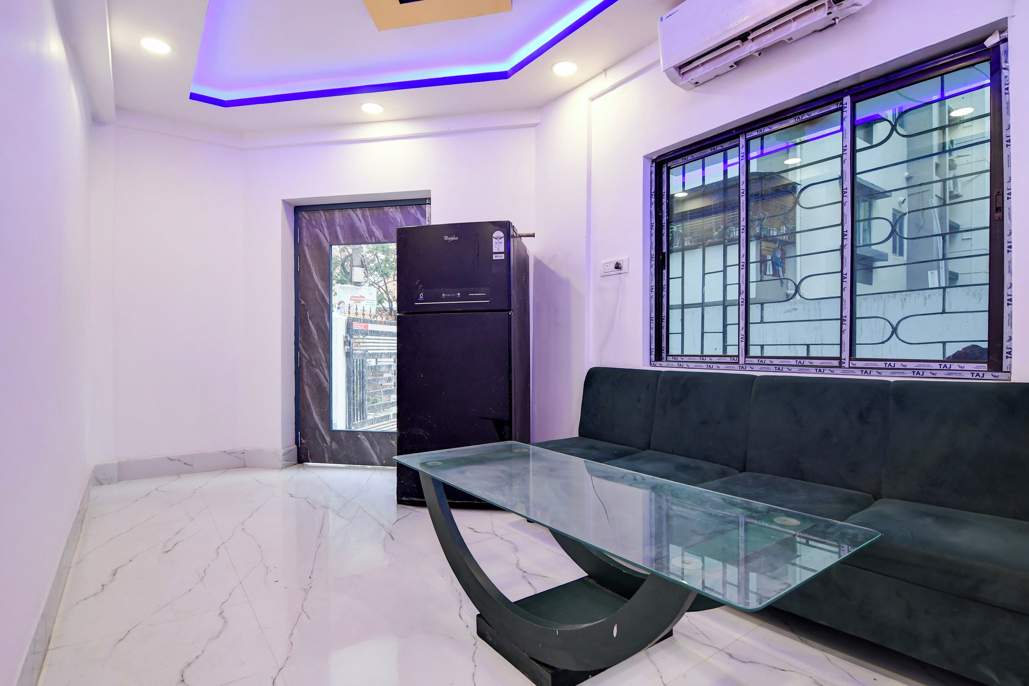 A modern living room with a glass coffee table, a black sofa, a refrigerator, and a window with a view of buildings.